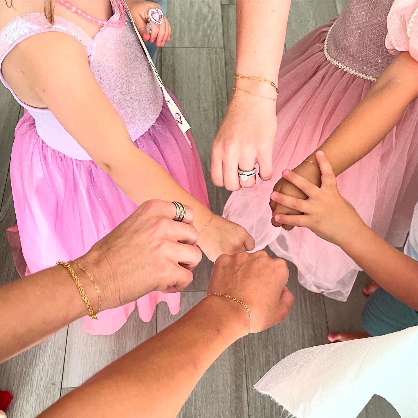 Overhead view of hands joined in a circle — two children in pink tulle princess dresses and two adults showing delicate gold bracelets and rings, a playful family moment.