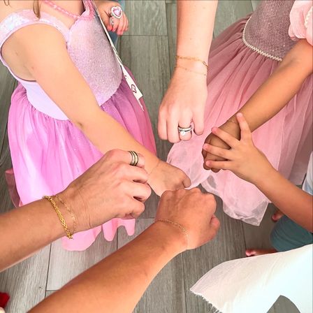 Overhead view of hands joined in a circle — two children in pink tulle princess dresses and two adults showing delicate gold bracelets and rings, a playful family moment.