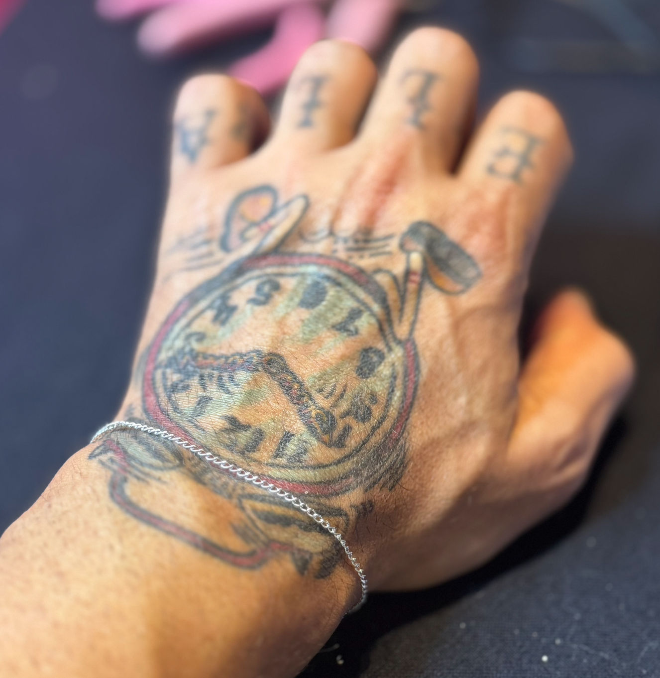 Close-up of a tattooed hand showing a vintage pocket-watch tattoo on the back of the hand and a delicate silver chain bracelet at the wrist, blurred dark background.