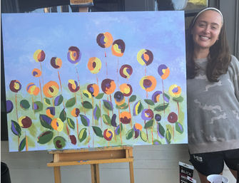 Smiling woman standing next to a large playful abstract floral painting on an easel, featuring circular yellow, orange and purple blooms with green leaves against a lavender sky backdrop.