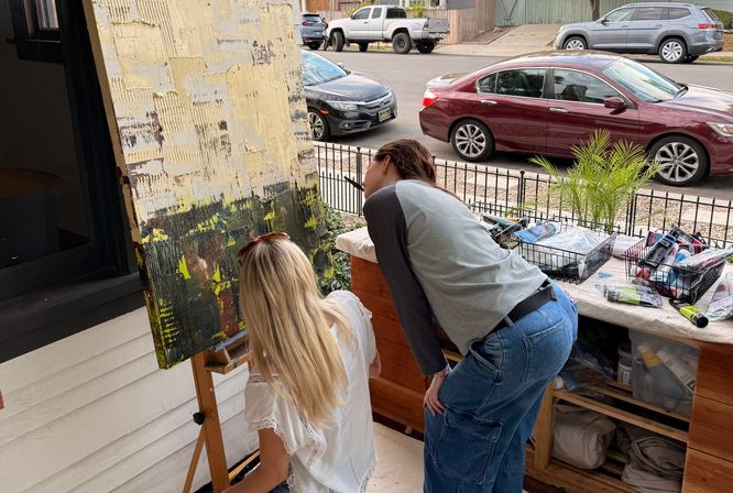 Two artists leaning over a textured abstract canvas on an easel at a suburban front porch, art supplies in baskets on the table and parked cars along a quiet residential street in the background