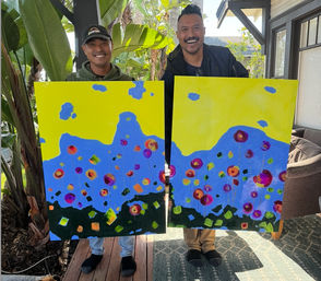 Two smiling people on a sunny tropical patio hold large, matching colorful abstract paintings of stylized flowers with bright yellow skies and blue hills