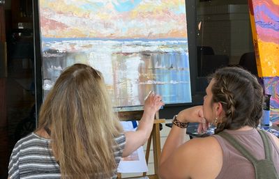 Two people seen from behind at an easel during a painting lesson, one guiding a textured pastel seascape canvas (acrylic)