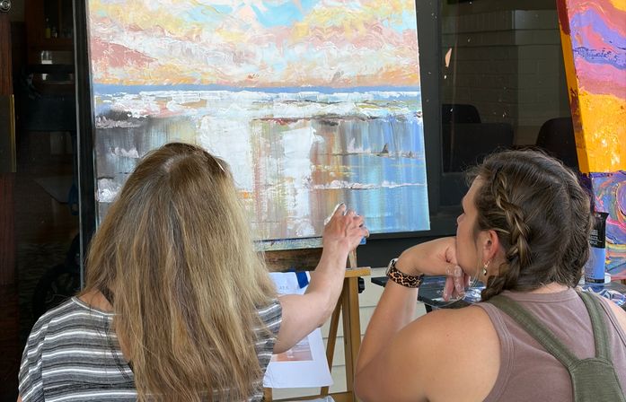 Two people seen from behind at an easel during a painting lesson, one guiding a textured pastel seascape canvas (acrylic)