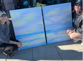 Two smiling people crouch on a sunny sidewalk outdoors beside two large pastel, beach‑inspired abstract seascape canvases with soft blue, pink and yellow cloud-like washes.