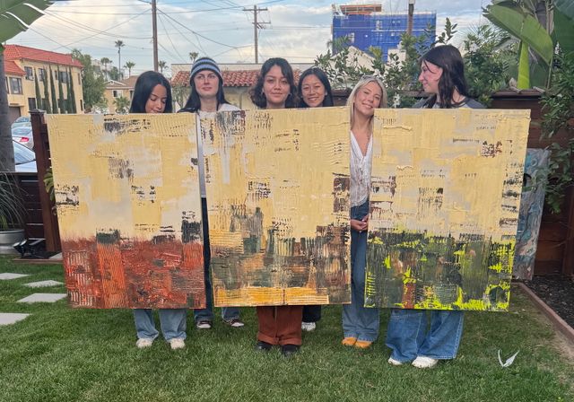 Six people stand behind three large textured abstract canvases in yellow and earthy tones, posed on grass in a palm-lined urban backyard with houses and utility wires in the background.