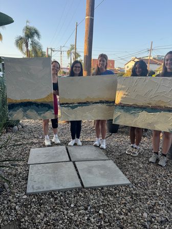 Group of five people at an outdoor art party holding large textured abstract canvases at golden hour, palm trees and utility poles in the background and a gravel path in the foreground.