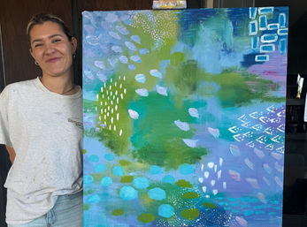 Smiling person standing beside a large colorful abstract canvas with teal, green, blue and purple washes, white dotted marks and geometric accents — contemporary studio artwork