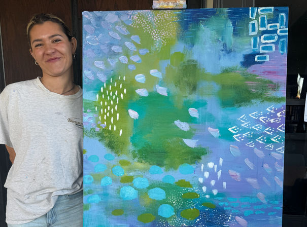Smiling person standing beside a large colorful abstract canvas with teal, green, blue and purple washes, white dotted marks and geometric accents — contemporary studio artwork