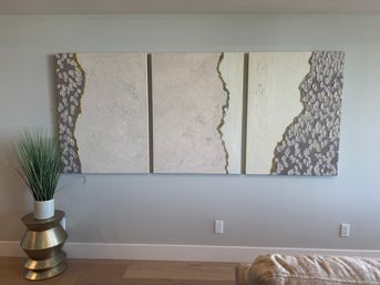 Modern living room wall featuring a three-panel abstract textured canvas triptych in neutral beige, cream and gray with gold accents, plus tall potted decorative grass on a stacked metallic side table and light hardwood floor.