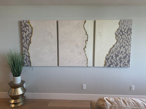Modern living room wall featuring a three-panel abstract textured canvas triptych in neutral beige, cream and gray with gold accents, plus tall potted decorative grass on a stacked metallic side table and light hardwood floor.