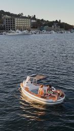 Three people lounging on a small teak-deck leisure boat near a busy marina and yachts along an urban waterfront shoreline in warm evening light