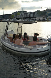 Six people relaxing in a small motorboat hot tub in a Seattle marina at sunset, with the Space Needle, waterfront buildings, and docked yachts in the background.
