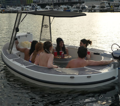 Six people relaxing in a small motorboat hot tub in a Seattle marina at sunset, with the Space Needle, waterfront buildings, and docked yachts in the background.