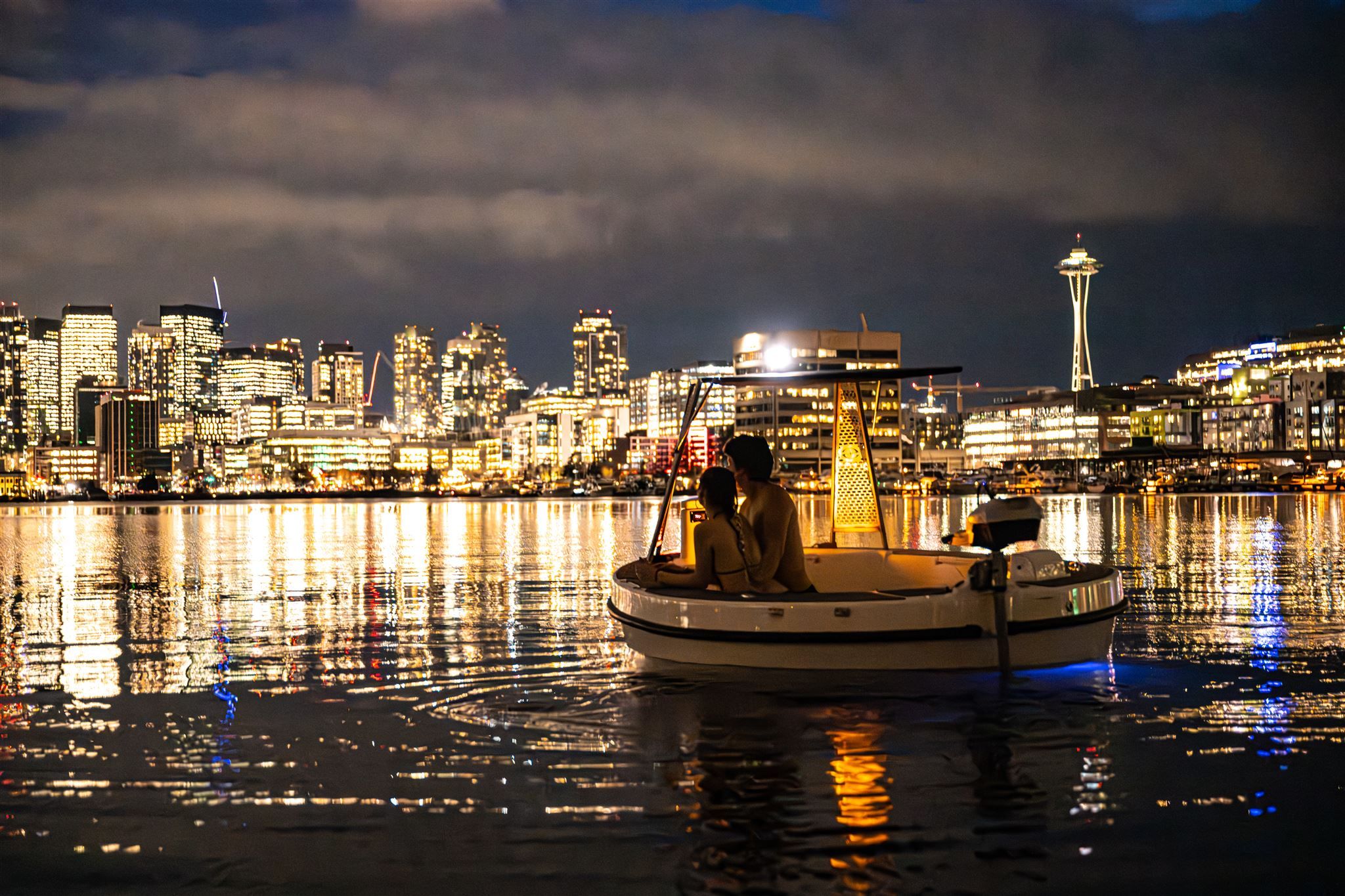 Couple on a cozy illuminated boat at night on calm water with the Seattle skyline and Space Needle glowing in the background, city lights sparkling and reflecting on the rippled water.