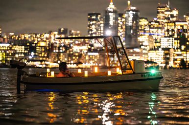 Candlelit small boat with a person relaxing on reflective water at night, blurred downtown city skyline glowing in the background.