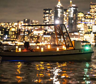 Candlelit small boat with a person relaxing on reflective water at night, blurred downtown city skyline glowing in the background.
