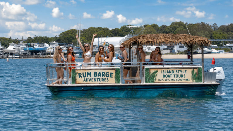 Group of women in swimsuits enjoying drinks on a tiki-style party boat tour in a sunny coastal marina with yachts and a sandy shoreline in the background
