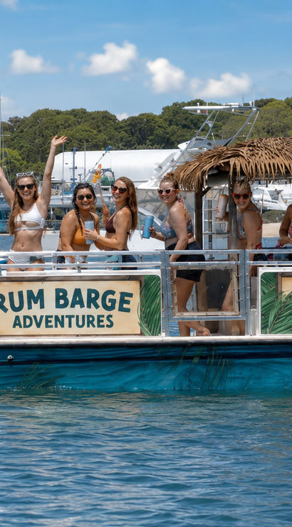 Group of women in swimsuits enjoying drinks on a tiki-style party boat tour in a sunny coastal marina with yachts and a sandy shoreline in the background
