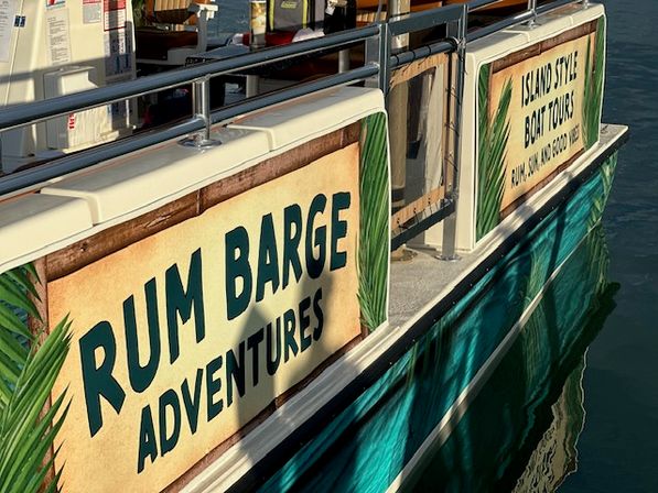 Sunlit rum-themed party boat docked at a marina with teal hull, tropical leaf signage advertising boat tours, and reflections in calm water.