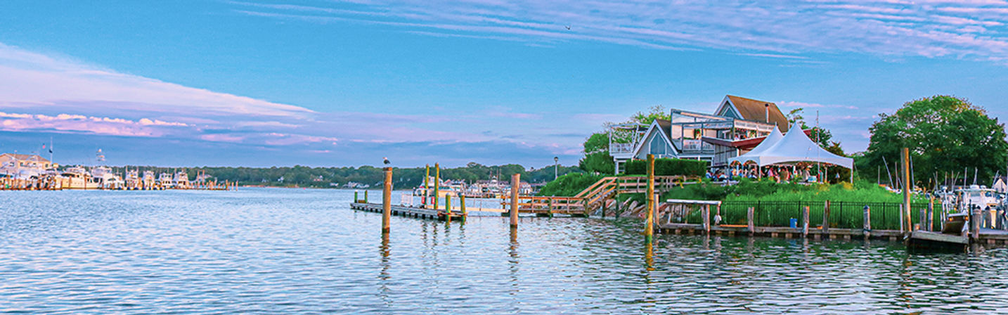 Coastal marina scene with waterfront dining under a white tent, wooden docks and moored boats on calm water beneath a pastel blue sky.