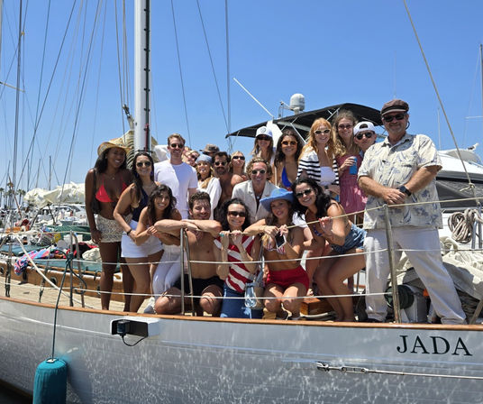 Large group of friends smiling and posing on the deck of a sailboat at a sunny marina — summer swimsuits, hats, sunglasses, and clear blue sky.