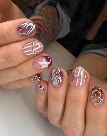 Close-up of hands showing trendy mixed nail art manicure — pink, burgundy and brown gel nails with white stripes, star and polka-dot designs, metallic silver 3D accents and marbled swirls, glossy finish, tattooed forearm in background.