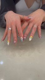 Close-up of hands showing trendy almond acrylic nails with white, yellow and blue swirl designs, tiny rhinestones and mirrored accents, paired with silver rings over a marble countertop.