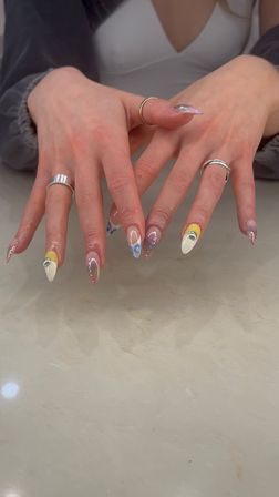 Close-up of hands showing trendy almond acrylic nails with white, yellow and blue swirl designs, tiny rhinestones and mirrored accents, paired with silver rings over a marble countertop.