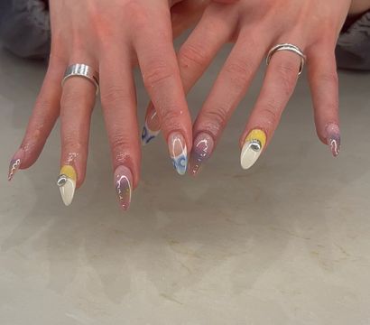 Close-up of hands showing trendy almond acrylic nails with white, yellow and blue swirl designs, tiny rhinestones and mirrored accents, paired with silver rings over a marble countertop.