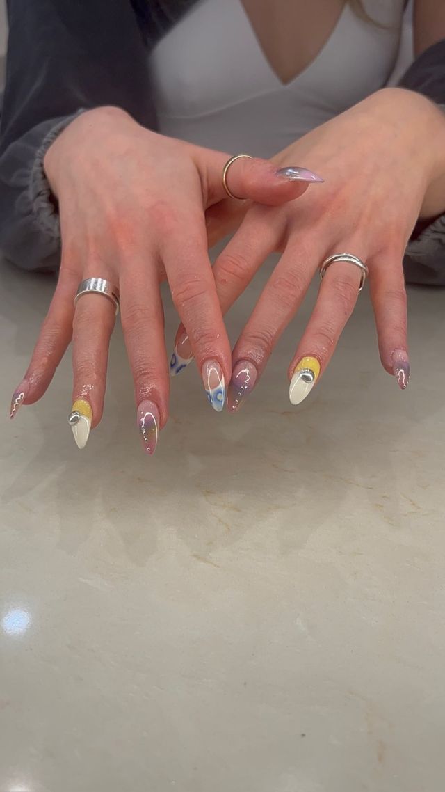 Close-up of hands showing trendy almond acrylic nails with white, yellow and blue swirl designs, tiny rhinestones and mirrored accents, paired with silver rings over a marble countertop.