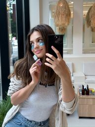 Smiling woman taking a mirror selfie in a bright salon, wearing blue under-eye patches and holding a small skincare bottle