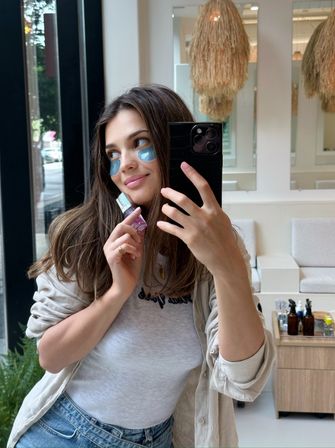 Smiling woman taking a mirror selfie in a bright salon, wearing blue under-eye patches and holding a small skincare bottle