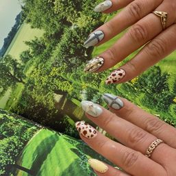 Almond-shaped manicure with mixed nail art — brown cow-print, pale blue geometric and studded designs — hand with gold rings over a vivid green countryside backdrop.