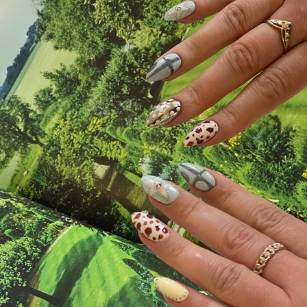 Almond-shaped manicure with mixed nail art — brown cow-print, pale blue geometric and studded designs — hand with gold rings over a vivid green countryside backdrop.