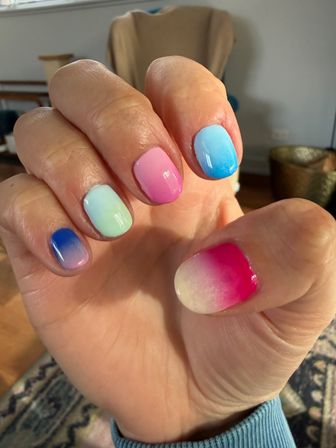 Close-up of a hand with short rounded glossy ombre manicure in pastel gradients — pink, blue, mint and purple — indoor living-room background