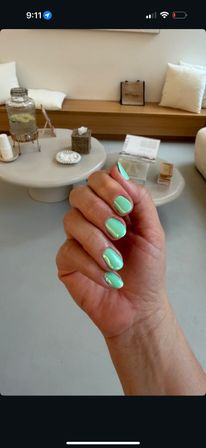 Close-up of a hand showing glossy mint-green oval manicure in a modern nail salon waiting area with minimalist white tables and cushions.