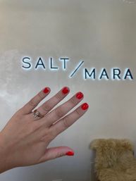 Close-up of a hand with glossy red manicure and stacked diamond rings held up in front of an illuminated wall sign in a modern nail salon interior.