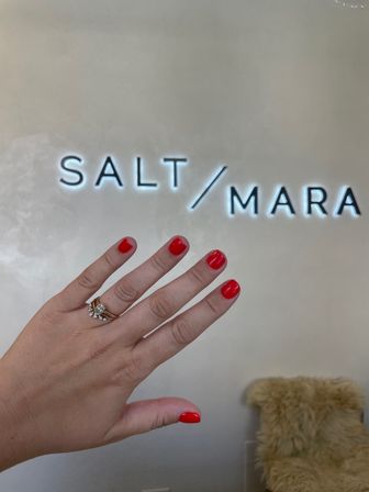 Close-up of a hand with glossy red manicure and stacked diamond rings held up in front of an illuminated wall sign in a modern nail salon interior.