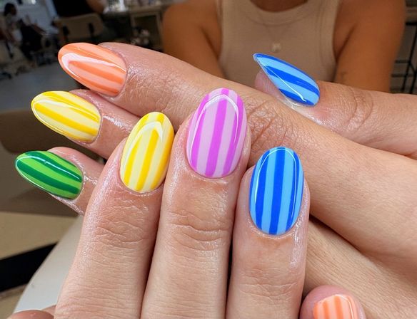 Close-up of glossy almond-shaped nails with bold vertical stripes in rainbow hues (green, yellow, orange, pink, blue) — trendy striped manicure.