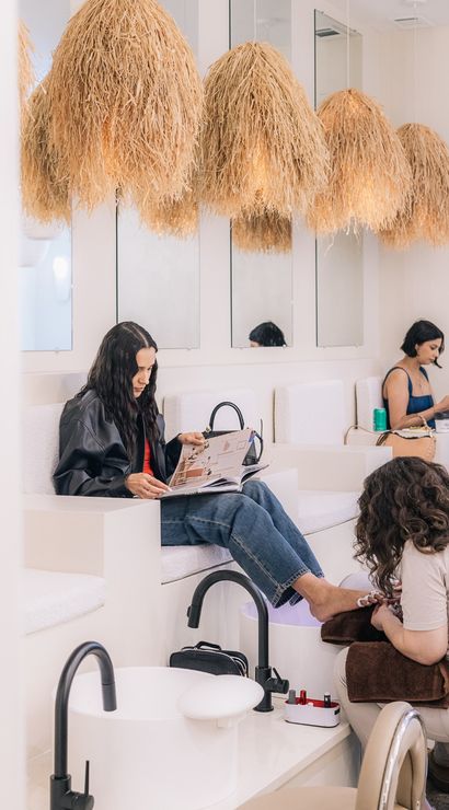 Bright minimalist nail salon interior with oversized straw pendant lights; a woman reads a magazine while getting a pedicure from a technician and another client scrolls on her phone.