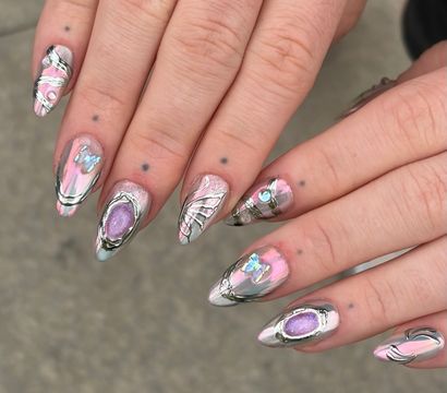 Close-up of hands displaying stiletto-shaped pink iridescent chrome nail art with silver 3D swirls and purple gem-like accents on a neutral gray background.
