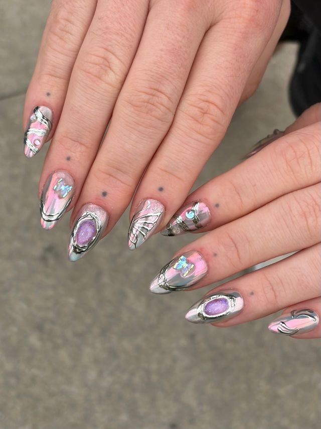 Close-up of hands displaying stiletto-shaped pink iridescent chrome nail art with silver 3D swirls and purple gem-like accents on a neutral gray background.