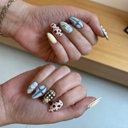 Close-up of hand with stiletto nail art and manicure in pastel blue and cream, featuring cow-print accents, silver studs, pearls and a cross design.