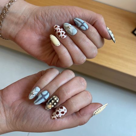Close-up of hand with stiletto nail art and manicure in pastel blue and cream, featuring cow-print accents, silver studs, pearls and a cross design.
