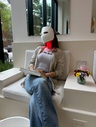 Person relaxing in a modern spa waiting area wearing a red-glowing LED phototherapy face mask, seated with a journal beside a small vase of flowers.