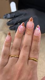 Glossy almond-shaped gel manicure with nude base and brown tortoiseshell French tips, delicate white-and-pink floral accents on two nails, gold rings on fingers and a gloved technician hand in the background – salon nail art close-up.