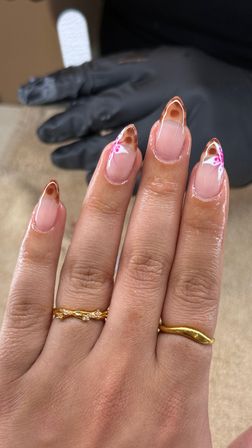Glossy almond-shaped gel manicure with nude base and brown tortoiseshell French tips, delicate white-and-pink floral accents on two nails, gold rings on fingers and a gloved technician hand in the background – salon nail art close-up.