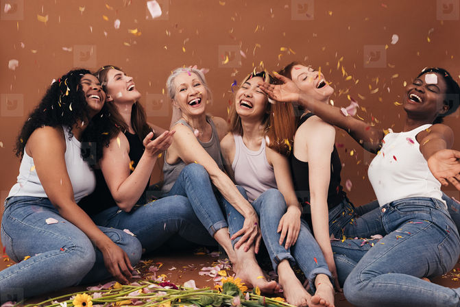 Six diverse women of different ages sitting on the floor in jeans and tank tops, laughing and celebrating as flower petals and confetti fall around them with scattered flowers on the floor.