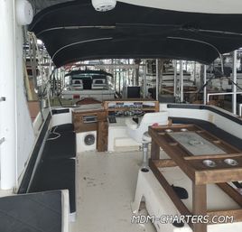 Cozy covered motorboat cockpit in a marina slip with black bimini top, wraparound bench seating, helm station, wooden table with cup holders and other docked boats visible in the background